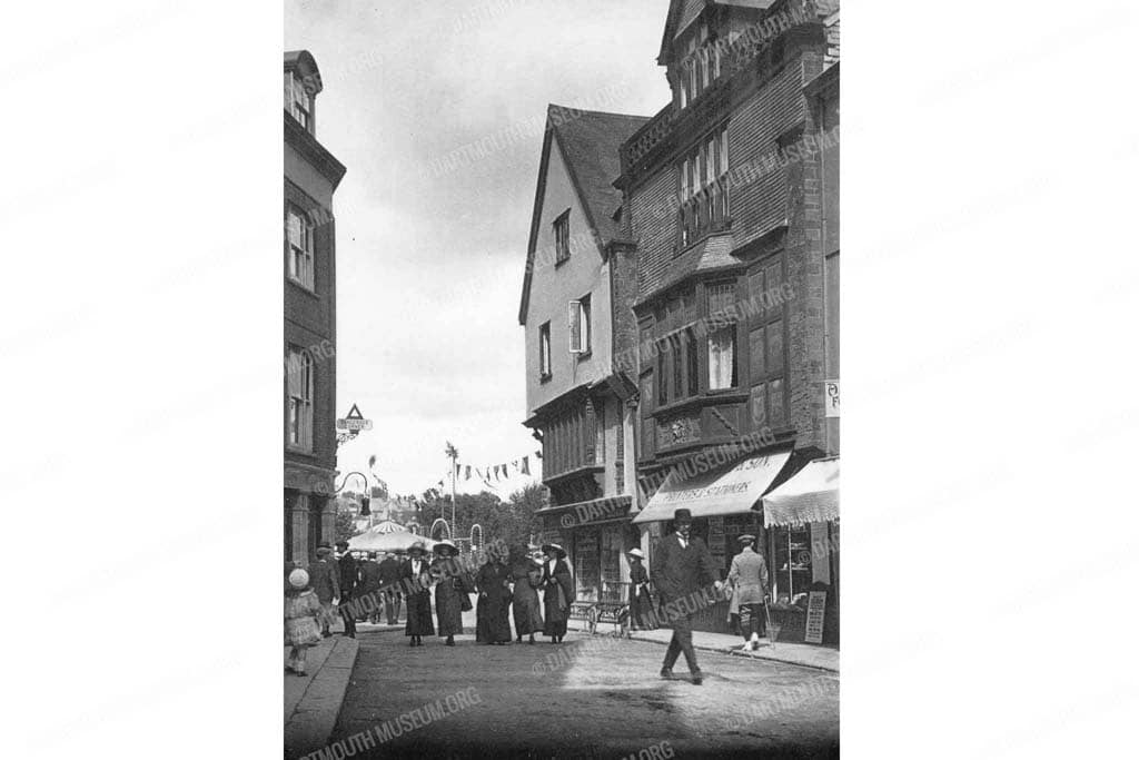 Historical photograph of Fairfax Place in Dartmouth looking towards the Quay between 1909 - 1914, showing a busy scene with happy people walking past the local supply stores.